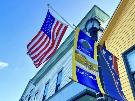 An American flag and a blue Welcome to Eudora street banner flying from a lamppost on Main Street, against a clear blue sky