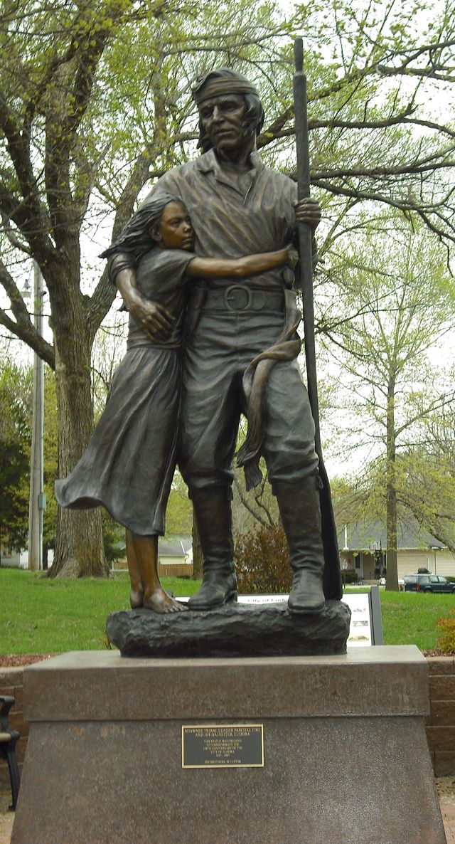 Bronze statue of Shawnee leader Paschal Fish with his daughter Eudora hugging his side, standing on a granite base in a tree-lined park